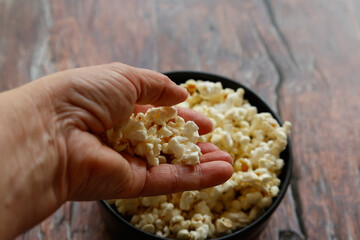 A close-up of a hand holding a handful of popcorn above a black bowl filled with popcorn. The rustic wooden table in the background adds a cozy and natural atmosphere to the scene.