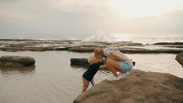 Happy young mother kissing cute son kid spending time together at stone beach sea wave slow motion