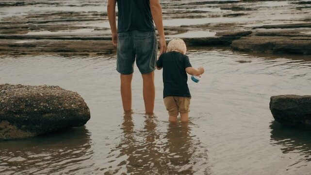 Father and son kid holding hands admiring sea landscape wave water stone rock beach slow motion