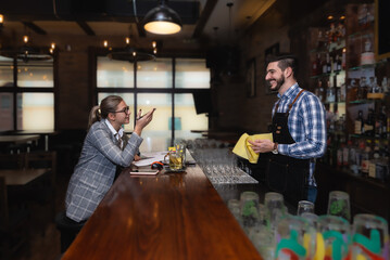 Satisfied restaurant owner checking monthly reports on laptop, bills and expenses of her small business. Start-up entrepreneur happy about financial reports while talking to the waiter or barman.