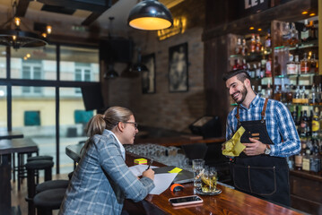 Satisfied restaurant owner checking monthly reports on laptop, bills and expenses of her small business. Start-up entrepreneur happy about financial reports while talking to the waiter or barman.
