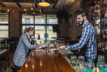 Satisfied restaurant owner checking monthly reports on laptop, bills and expenses of her small business. Start-up entrepreneur happy about financial reports while talking to the waiter or barman.