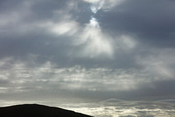 Cloudy sky with sun rays piercing through, silhouetted mountain in the foreground.  A gray and somber landscape.