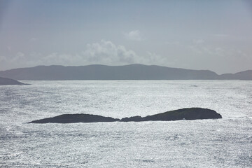 Fototapeta premium Ocean vista: Two small islands, silhouetted against a hazy, overcast sky, are visible on a sparkling, sunlit sea.