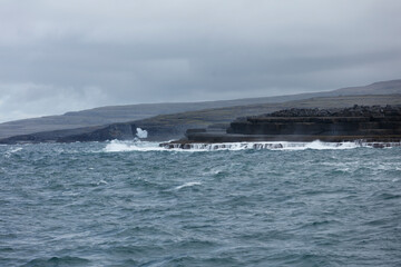 Rough seas crash against a dramatic, dark rock coastline under a gray sky.  Waves churn over tiered basalt formations.