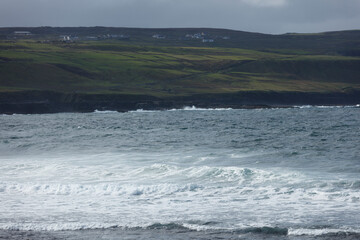 Ocean waves crash on a rocky coast, with a hillside of green fields and small buildings in the background.  Coastal landscape.