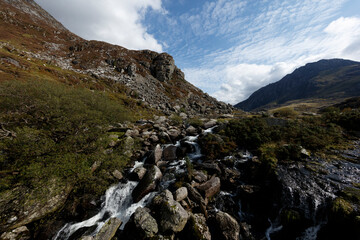 Mountainous landscape with a cascading stream.  Rocky terrain, vegetation, and a clear blue sky are prominent.