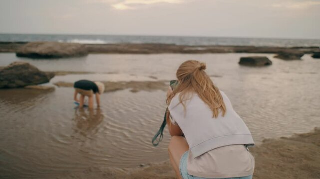 Caring mother shooting video baby son playing ship toy floating sea beach seascape use smartphone
