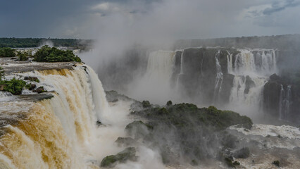 An impressive waterfall landscape. Streams of bubbling white water fall from the ledge into the abyss. Spray and fog rise into the sky. green vegetation in the riverbed. Iguazu Falls. Brazil.