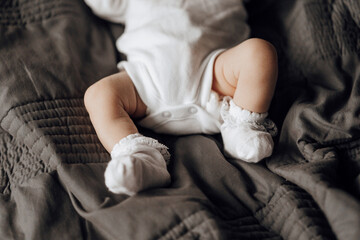 A baby's legs are shown in white socks on a bed. The baby is wearing a white onesie and he is sleeping