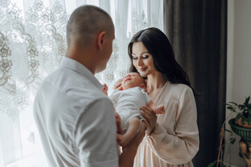A man and a woman are holding a baby in front of a window. The baby is smiling and the family is happy