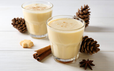 Tasty Christmas cocktail (eggnog) in glasses, pine cones and spices on white wooden table, closeup