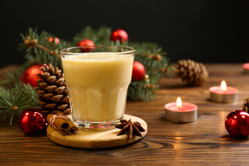 Tasty Christmas cocktail (eggnog) in glass, festive decor and burning candles on wooden table, closeup