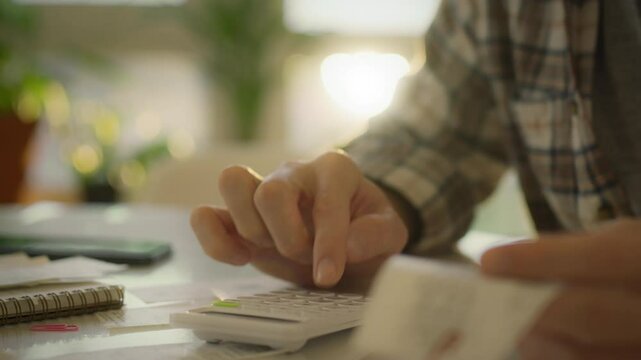 A person is using a calculator while reviewing financial documents in a warm and inviting workspace filled with natural light. Papers and a notebook are scattered on the table nearby