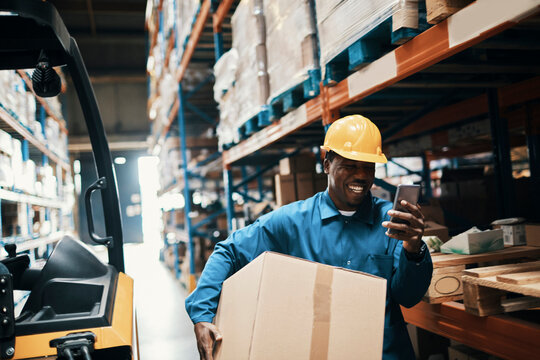 Warehouse worker with hard hat using smartphone while carrying box