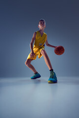 Low angle view of teenage boy, dressed vibrant basketball gear, training dribbling technique in action against grey studio background. Concept of sport, leisure activity, hobby, competition. Ad