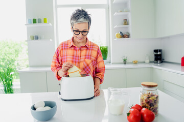 Elderly woman in casual style preparing breakfast in a bright kitchen, adding a slice of bread to...