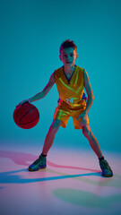 Sportive teenage boy, basketball player, dressed yellow-blue uniform, playing game in neon light against gradient studio background. Concept of sport, leisure activity, hobby, competition. Ad