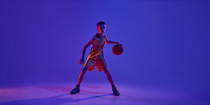 Confident, teenage ball stands ready to start game and get victory in this decisive basketball match in neon light against gradient studio background. Concept of sport, leisure activity, competition.