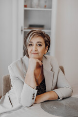 A woman in a gray suit is sitting at a table with her hands on her knees. She is smiling and she is in a good mood