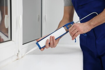 Worker with caulking gun sealing window indoors, closeup