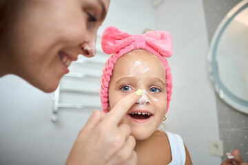 Smiling young woman and little daughter with a cosmetic bow on their heads smearing face cream on...