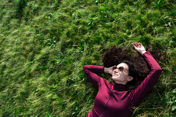 Curly haired woman wearing sunglasses lying down on a grass. Attractive cheerful lady relaxing outdoors. Copyspace