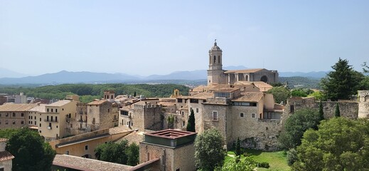 Naklejka premium A panoramic view of the medieval town of Girona, Spain. The historic buildings, including the Girona Cathedral, are nestled amidst rolling hills and a clear blue sky.