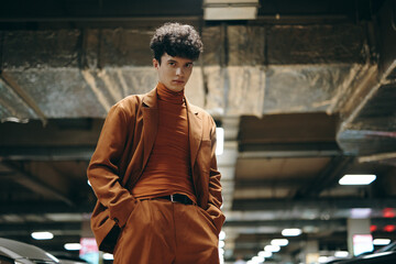 A stylish young man in an orange suit and turtleneck poses confidently in a dimly lit parking garage