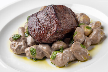 A gourmet dish featuring a perfectly seared piece of beef on a bed of creamy mushroom gnocchi, garnished with vibrant green pesto and drizzled with olive oil.