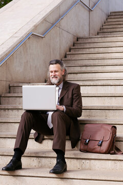 positive mature male entrepreneur in elegant suit siting on urban stairway and working online on laptop in city, concept of business and middle age man lifestyle