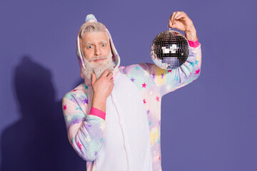Cheerful mature man in unicorn pajama holding a disco ball against a purple background