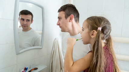 Fototapeta premium A loving father is holding his daughter in the arms and helping her brushing their teeth in a bathroom.