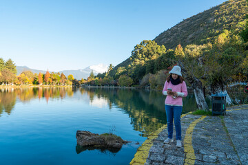 Asian female tourist holding a smartphone relaxes in the morning while visiting Black Dragon Pool, Lijiang Park, China.
