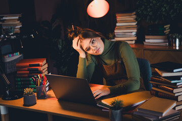 Young woman in casual green jumper studying late at night with laptop and books, embodying student...