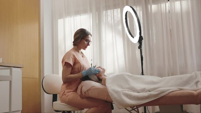A therapist in professional attire performs a lymphatic drainage massage in a serene spa setting with a ring light and a relaxed patient.