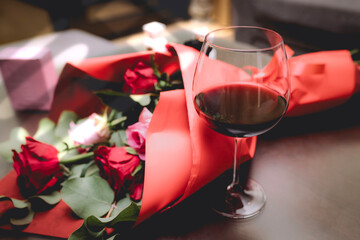 A glass of red wine placed beside a bouquet of red and pink roses wrapped in vibrant red paper, creating a romantic and elegant setting.