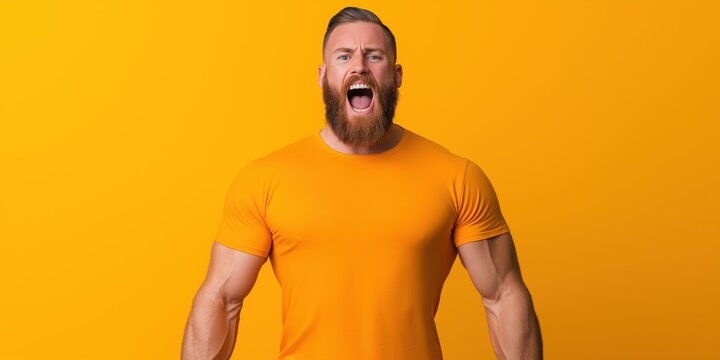 A man with a beard and orange shirt is standing in front of a yellow wall. He is making a loud noise, which could be interpreted as yelling or shouting. Concept of energy and excitement