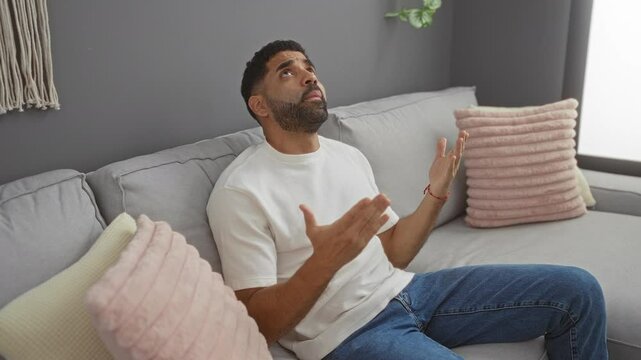 Young man in a white shirt sitting on a sofa in a living room covers his ears with hands due to noise from upstairs, looking frustrated and distressed.