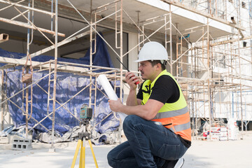 Male construction engineer worker working with blueprint and using surveyor's telescope or level measuring instrument at construction site