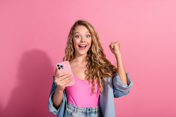 Joyful young woman in stylish casual denim celebrating success with a smartphone against a vibrant pink background