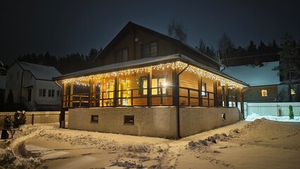 Obraz premium Garlands are lit on the walls and terrace of a wooden cottage in the evening darkness on Christmas Eve. Everything around is covered in snow. The path is cleared in snowdrifts. Nearby is a forest