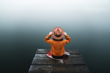 Back view of fashioned young woman sitting on wooden dock looking at view on a misty morning. Female hipster with brown hat relaxes on the edge of jetty admiring foggy lake. Wonderful nature getaway
