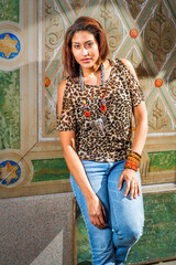 A woman leans against a decorative wall, wearing a leopard print top and jeans. Bold jewelry with multiple bracelets and a statement necklace enhances her fashionable look.