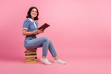 Smiling young woman sitting on stack of books holding a book against pink background. Casual style with striped knitwear and jeans.