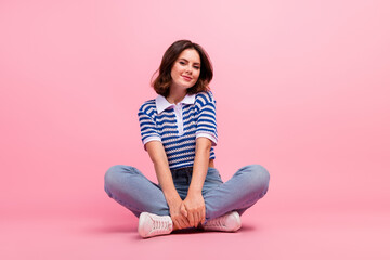 Charming young woman in stylish striped t-shirt and jeans sitting cross-legged against pink background