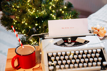 Typed on a typewriter: Happy New Year 2025, against the backdrop of a Christmas tree with garland and cocoa with tangerines on a wooden tray