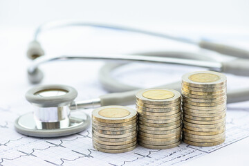 Healthcare economics : The cost of care and financial planning in medicine. A stethoscope resting beside stacks of coins on an electrocardiogram graph, depicting concepts of medical expenses, etc