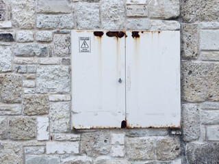 A white-painted electrical panel with rust stains adds industrial contrast to a historical stone wall. The aged textures create a unique blend of modern utility and antique architecture.