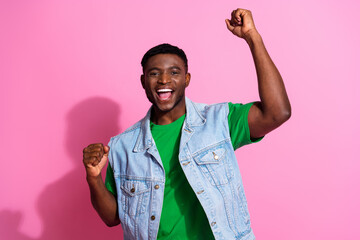 Cheerful young man with raised fists celebrating success, wearing denim vest and green shirt against vibrant pink background